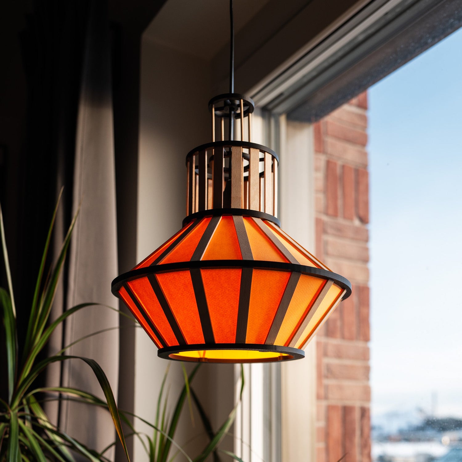 Hängande design-lampskärm i gult med svarta delar och trädetaljer, lyser i ett soligt fönster